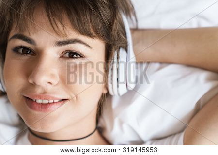Photo of a cheerful optimistic young beautiful woman lies indoors at home on bed listening music with headphones.