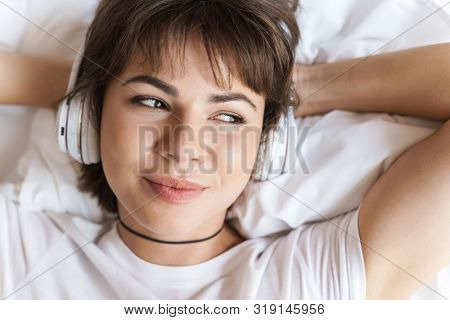 Photo of a dreaming thinking optimistic positive pleased young beautiful woman lies indoors at home on bed listening music with headphones looking aside.