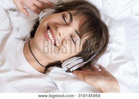 Photo of a optimistic positive cheery young beautiful woman lies indoors at home on bed listening music with headphones.
