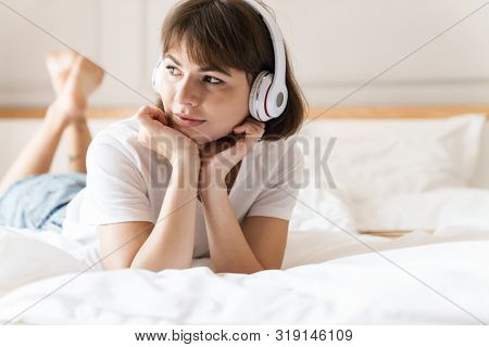 Photo of a happy optimistic positive pleased young beautiful woman lies indoors at home on bed listening music with headphones.