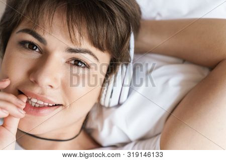 Photo of a optimistic positive cheery young beautiful woman lies indoors at home on bed listening music with headphones.