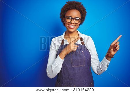 Young african american woman shop owner wearing business apron over blue background smiling and looking at the camera pointing with two hands and fingers to the side.