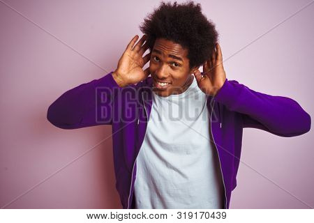 Young african american man wearing purple sweatshirt standing over isolated pink background Trying to hear both hands on ear gesture, curious for gossip. Hearing problem, deaf