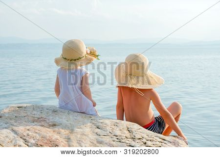Back View Of Two Kids Sitting On Stone And Looking To Ocean. Little Travelers Near The Ocean. Boy An