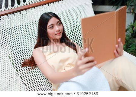 Young And Cute Asian Girl Lying In A Hammock And Reading A Book During Holiday