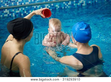 Front View Of Baby Boy During Swimming Lesson. Back View Of Mother Holding Child And Woman Pouring W