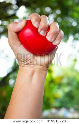 Woman's Left Hand Squeezing Red Stress Ball On Bokeh Green Garden Background, Exercise And Massage C