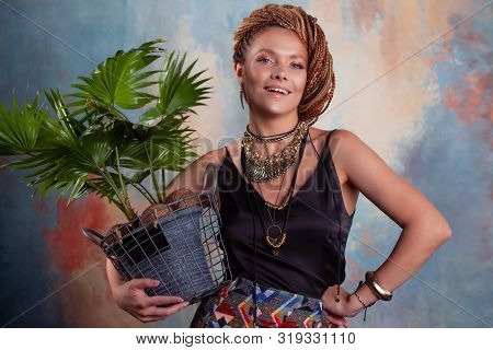 Southern Flavor. A Young Tanned Woman With Afro Braids Smiles Holding A Big Pot With A Tropical Plan