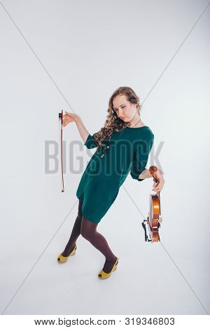 A Broken Doll In A Green Dress With A Violin And A Bow In Her Hands On A White Background. Tired Gir