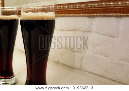 Two Glasses Poured With Dark Beer Or Soft Drink With Foam Head. Close Up Image Against White Brick W
