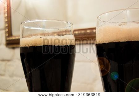 Two Glasses Poured With Dark Beer Or Soft Drink With Foam Head. Close Up Image Against White Brick W
