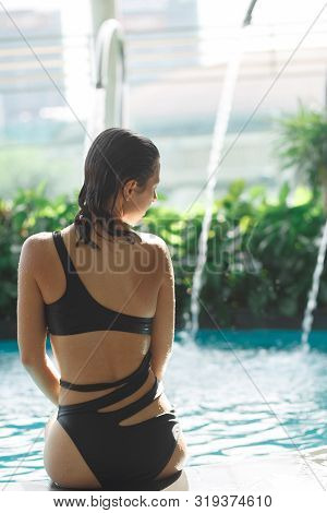 Shoot From Back Of Sexy Slim Female In Swimsuit Sits On Edge Of Swimming Pool Between Green Bushes O