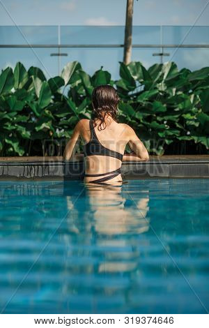 Shoot From Back Of Sexy Slim Female In Swimsuit Sits On Edge Of Swimming Pool Between Green Bushes O