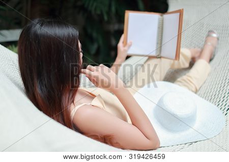 Young And Cute Asian Girl Lying In A Hammock And Reading A Book During Holiday