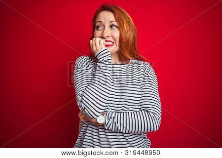 Young redhead woman wearing strapes navy shirt standing over red isolated background looking stressed and nervous with hands on mouth biting nails. Anxiety problem.