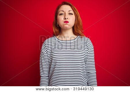 Young redhead woman wearing strapes navy shirt standing over red isolated background puffing cheeks with funny face. Mouth inflated with air, crazy expression.
