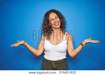 Middle age senior woman with curly hair standing over blue isolated background smiling showing both hands open palms, presenting and advertising comparison and balance