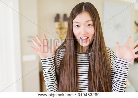 Beautiful Asian woman wearing stripes sweater over living room background celebrating crazy and amazed for success with arms raised and open eyes screaming excited. Winner concept
