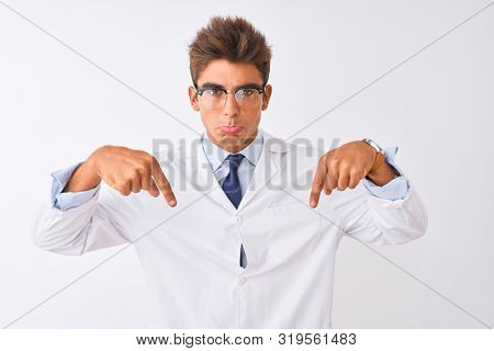 Young handsome sciencist man wearing glasses and coat over isolated white background Pointing down looking sad and upset, indicating direction with fingers, unhappy and depressed.