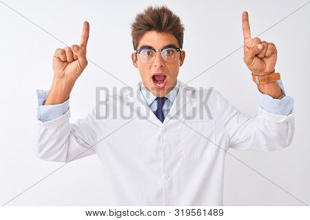 Young handsome sciencist man wearing glasses and coat over isolated white background smiling amazed and surprised and pointing up with fingers and raised arms.
