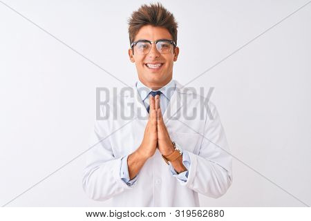 Young handsome sciencist man wearing glasses and coat over isolated white background praying with hands together asking for forgiveness smiling confident.