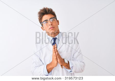 Young handsome sciencist man wearing glasses and coat over isolated white background begging and praying with hands together with hope expression on face very emotional and worried. 
