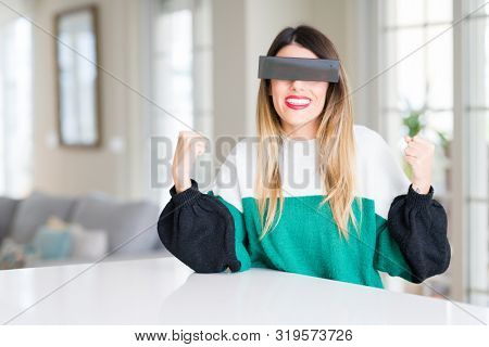 Young beautiful woman blindfold eyes with black censor band at home celebrating surprised and amazed for success with arms raised and open eyes. Winner concept.