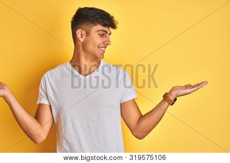 Young indian man wearing white t-shirt standing over isolated yellow background smiling showing both hands open palms, presenting and advertising comparison and balance