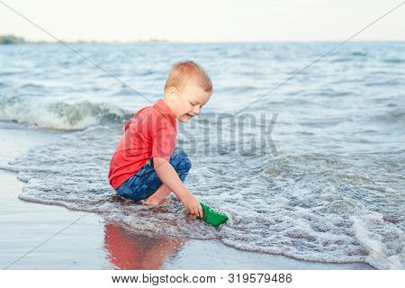 Happy Caucasian Red-haired Toddler Child Boy Putting Green Paper Boat In Water On Lake Sea Ocean Sho