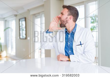Handsome doctor man wearing medical coat at the clinic shouting and screaming loud to side with hand on mouth. Communication concept.
