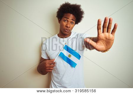 Afro american tourist man holding Argentinian Argentina flag over isolated white background with open hand doing stop sign with serious and confident expression, defense gesture