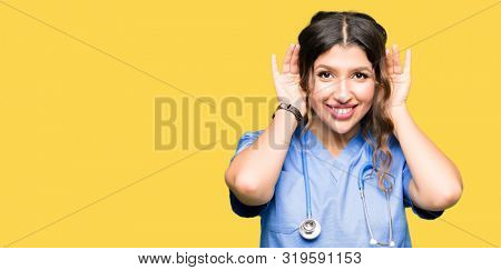 Young adult doctor woman wearing medical uniform Trying to hear both hands on ear gesture, curious for gossip. Hearing problem, deaf