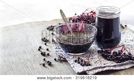Homemade Black Elderberry Syrup In Glass Bowl And Jar And Bunches Of Black Elderberry In Background.