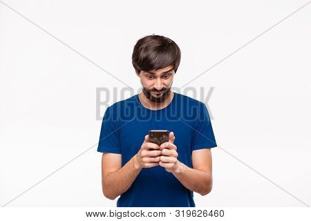 Good Looking Brunet Man In A Blue Shirt With Beard And Mostaches Shocked Holding Smartphone Looking 