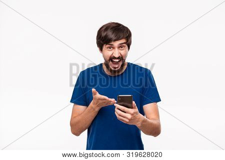 Good Looking Brunet Man In A Blue Shirt With Beard And Mostaches Shocked Holding Smartphone Looking 