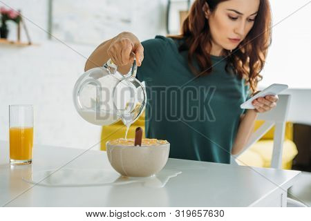 Woman Overfilling Bowl Of Flakes With Water While Sitting At Kitchen Table And Using Smartphone