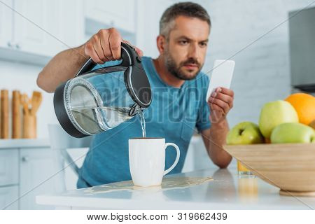 Selective Focus Of Man Overfilling Cup With Water While Sitting At Kitchen Table And Using Smartphon