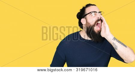 Young hipster man with long hair and beard wearing glasses shouting and screaming loud to side with hand on mouth. Communication concept.