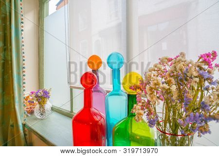 Mixed Colorful Bottles And Flowers On Window Sill Bright Red, Blue, Green And Orange.