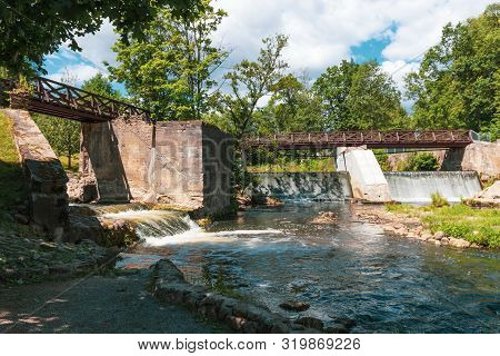Wooden Bridge Over The River Vilia In Vilnius With A Waterfall In The Park Belmonto.