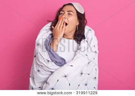 Close Up Portrait Of Young Woman In Sleep Mask Stands Yawning, Covering Her Mouth With Hand, Wearing