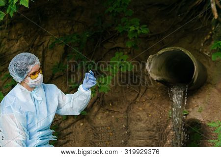 Female Ecologist Or Epidemiologist In A Protective Gown And Mask Visually Evaluates Test Tube Conten