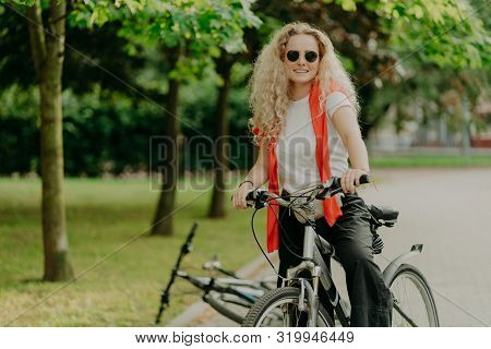 Glad Active Curly Haired Female Bicyclist Covers Distance On Her Own Transport, Wears Sunglasses, Ca