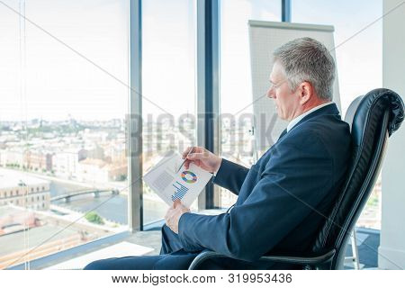 Mature Businessman Sitting Alone In Office With Large Panoramic Windows With View At City And Lookin