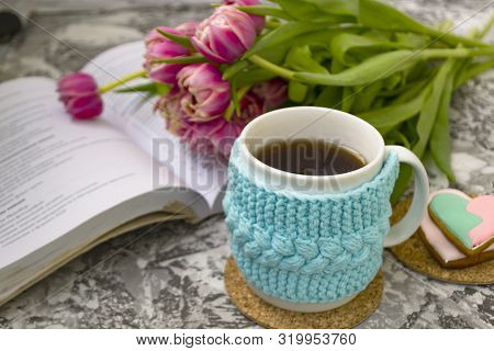 White Cup Of Tea In A Knitted Blue Case With An Open Book And With Pink Flowers Tulips And Sweets, W