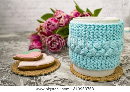 White Cup Of Tea In A Knitted Blue Case With Pink Flowers Tulips And Sweets, With Gingerbread On A T