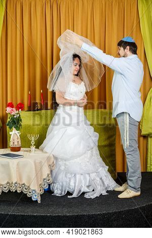 During The Chuppah Ceremony At A Synagogue Wedding, The Groom Lifts The Veil From The Bride's Face. 