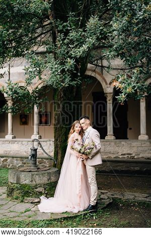 Groom Embraces Bride With A Bouquet Of Flowers In A Green Garden Against The Backdrop Of A Water Sta