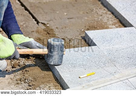 The Hands Of A Working Bricklayer Hold A Rubber Hammer And Lay Granite Paving Slabs On A Sandy Base 