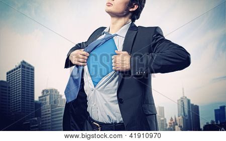 Image of young businessman showing superhero suit underneath his shirt standing against city background
